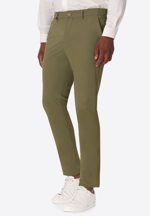 Uomo che indossa pantaloni slim fit verde oliva, sneakers bianche e camicia bianca a maniche lunghe, in piedi davanti a uno sfondo semplice.