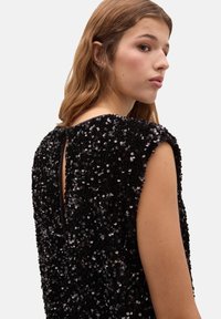 Femme aux cheveux châtain clair portant une robe noire sans manches ornée de sequins, avec une petite ouverture dans le dos et une fermeture à bouton au niveau du cou.