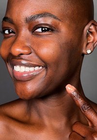 Kalju henkilö, jolla on sileä, tumma iho, hymyilee samalla kun osoittaa poskeaan. Hänellä on pienet korvakorut ja neutraali kynsilakka yhdellä sormella.