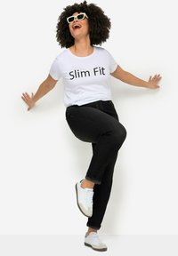 T-shirt bianca con la scritta "Slim Fit", jeans neri slim-fit, e sneakers bianche con dettagli grigi, modello in posa con le braccia allargate.