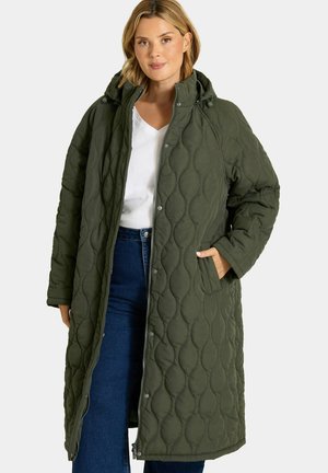Femme portant un long manteau matelassé vert foncé à capuche par-dessus un top blanc et un jean bleu, avec une main dans la poche du manteau.