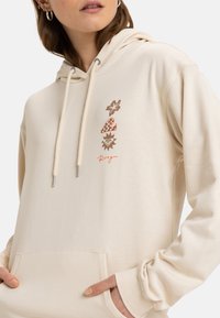 Crème hoodie met een voorzak, voorzien van bloem- en zongraphics in bruin en oranje. Verhoogde trekkoorden met metalen punten.