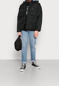 Musta hupparillinen takki, jossa kaksi suurta etutaskua, yhdistettynä mustaan t-paitaan, vaaleansinisiin farkkuihin ja mustavalkoisiin tennareihin.