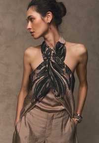 Woman with hair in a bun wears a sleeveless beige top with black textured patterns and matching pants, hands in pockets, looking left.