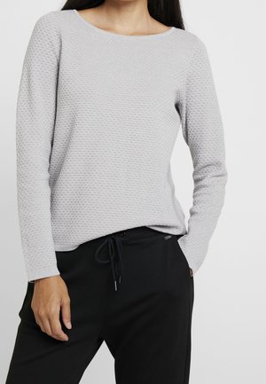 Femme portant un pull à manches longues texturé gris clair et un pantalon noir avec cordon, une main dans la poche, sur un fond uni.