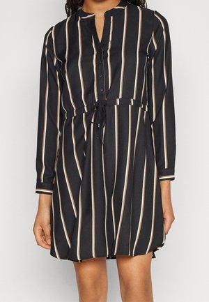 Zwarte lange mouwen jurk met verticale beige en witte strepen, knoopsluiting aan de voorkant en een ceintuur in de taille, gedragen door een staand persoon.
