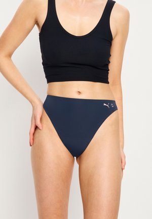 Femme portant un bas de bikini bleu marine taille haute Puma et un débardeur court noir, debout avec une main posée sur la hanche.