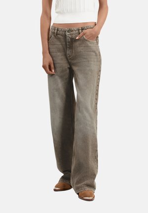 Jean large en denim vert olive délavé avec une texture subtile, cinq poches et une fermeture à bouton ; porté avec un haut blanc ajusté.