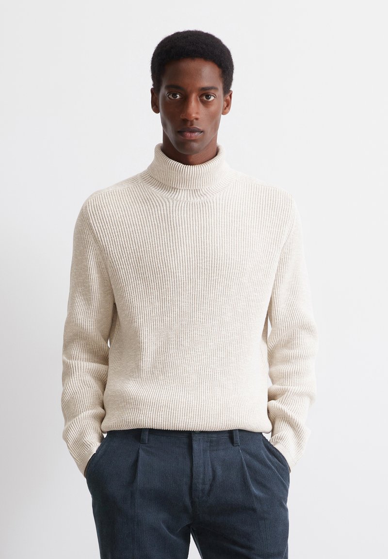 Cremfarbener, gerippter Rollkragenpullover aus strukturiertem Strickmaterial, kombiniert mit dunklen, gefalteten Hosen. Das Design hat einen hohen Kragen und lange Ärmel.