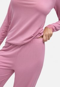 Top rosa de manga larga con borde de encaje y pantalones a juego hechos de un tejido suave y elástico. La parte superior cae holgadamente sobre la cinturilla de los pantalones.