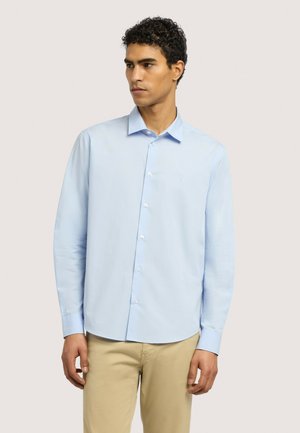 Mann trägt ein hellblaues Hemd mit klassischem Kragen und beige Hose, steht vor einem einfarbigen Hintergrund.