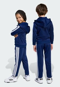 Námořnický sportovní komplet s bílými bočními pruhy, obsahující zipovou mikinu s kapucí a odpovídající kalhoty. Boty jsou bílé s černými akcenty.