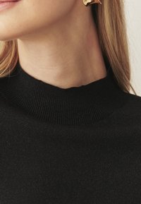 Schwarzer gestrickter Pullover mit hohem Kragen, der eine gerippte Textur und einen leichten Glanz aufweist, ergänzt durch sichtbare Aussparungen am Ausschnitt.