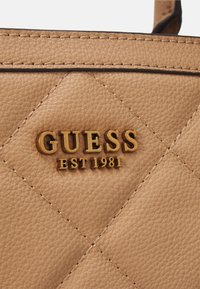 Borsa in pelle tan con motivo a diamante trapuntato, logo dorato "GUESS EST 1981" e cuciture a contrasto. Texture liscia, design strutturato.