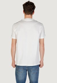 T-shirt bianco in cotone con maniche corte, caratterizzato da rifiniture nere lungo le cuciture delle spalle. Il retro è liscio con un taglio dritto all'orlo.