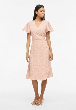 Mujer con un vestido cruzado floral color rosa claro con mangas cortas vaporosas y sandalias blancas de tacón con tiras, de pie y mirando hacia la derecha.