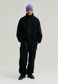 Czarna polarowa kurtka z dwoma zasuwanymi kieszeniami i czarne polarowe spodnie. Fioletowy prążkowany czapka z logo, noszona z czarnymi sznurowanymi butami.