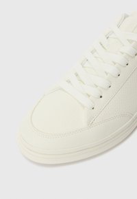 White leather sneaker with textured upper, round toe, and flat laces. Features understated stitching and a smooth rubber sole.