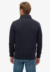 Marineblauwe sweatshirt met een hoge kraag, ribbelige onderrand en mouwen, en een gladde textuur. Heeft een kleine lus aan de achterkant van de hals voor ophangen.
