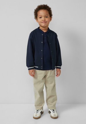 Małe dziecko stojące i uśmiechające się, ubrane w granatową kurtkę i polówkę, beżowe spodnie oraz białe trampki na jednolitym tle.