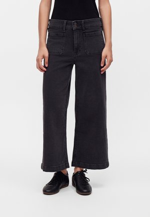 Person iført højtaljede, vidde sorte jeans med forlommer og sorte snøresko, stående mod en ensfarvet baggrund.