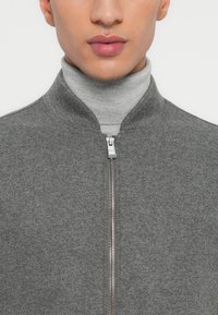 Primo piano di una persona che indossa un maglione grigio con zip sopra un dolcevita grigio chiaro, mostrando la parte inferiore del viso e la parte superiore del torso.