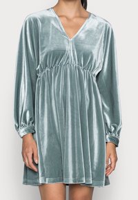 Sametové šaty v tlumené teal barvě s výstřihem do V, dlouhými nabíranými rukávy a elastickým pasem pro efekt shromážděné siluety.