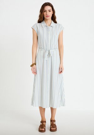 Kobieta w białej, bezrękawnikowej sukience sięgającej kostek z pionowymi niebieskimi paskami, zapinanej z przodu na guziki, z paskiem wiązanym w talii, w brązowych sandałach i z bransoletką.