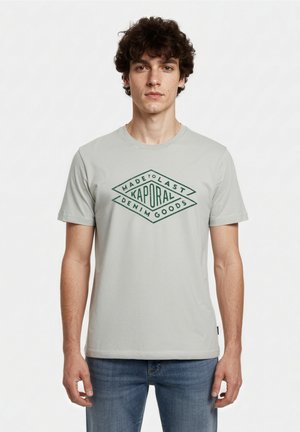 Joven con pelo rizado que lleva una camiseta gris claro con el logotipo "Kaporal Made to Last Denim Goods" y vaqueros azules.