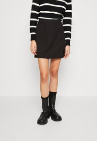 Mini-jupe noire au tissu lisse et avec des fentes sur les côtés, associée à des bottines noires brillantes et un pull rayé noir et blanc.