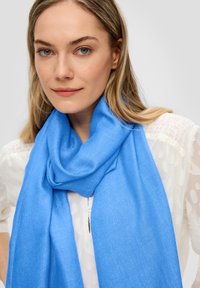 Foulard bleu clair en tissu doux, avec une texture lisse et un design drapé, porté sur un chemisier blanc texturé.