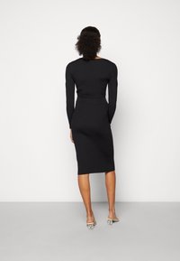 Frau mit lockigem Haar, die ein schwarzes geripptes langärmliges Midikleid und beige Mules mit durchsichtigen runden Absätzen trägt, steht vor einer weißen Wand.