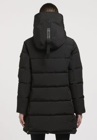 Persona, tērpusies garā melnā pūkainajā mētelī ar lielu pievienotu kapuci un redzamu zīmola jostu aizmugurē, stāv pretī vienkāršai fona.