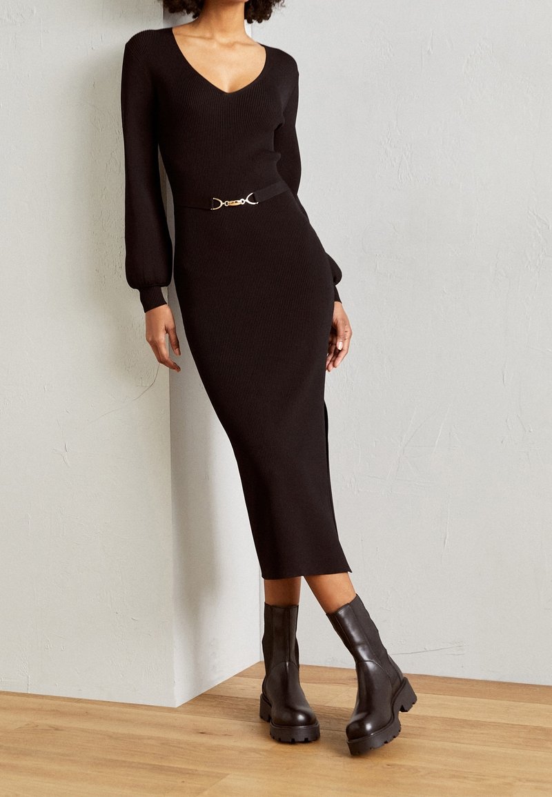 Femme portant une robe midi noire à manches longues avec ceinture et des bottines noires à semelle épaisse, debout devant un mur clair et un sol en bois.