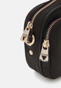 Sort nylon taske med guld hardware, udstyret med dobbelte lynlåse og fremtrædende 'GUESS' branding på lynlåsene. Glat tekstur, kompakt form.