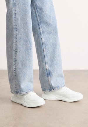 Personne portant un jean bleu clair à jambes larges et des baskets blanches épaisses, debout sur un sol gris contre un mur blanc.