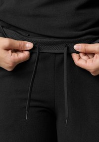 Schwarze Fleece-Hosen mit einem strukturierten Bund und verstellbaren Kordeln. Nahaufnahme von Händen, die den Bund zur Anpassung halten.