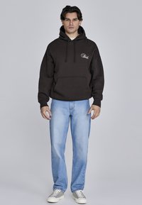 Sudadera con capucha marrón y bolsillo frontal, combinada con vaqueros rectos de color azul claro y zapatillas grises. La sudadera cuenta con un logo bordado en blanco.