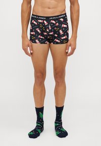 Schwarze Boxershorts mit einem Weihnachtsmann-Muster, ausgestattet mit einem weichen Bund mit "JACK & JONES" Logo. Kombiniert mit dunklen Socken, die Weihnachtsbäume zeigen.
