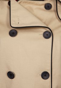 Beige trenchcoat stof met zwarte piping, met een dubbel geknoopt ontwerp en donkere knopen. Glad textuur met zichtbare stiksels.