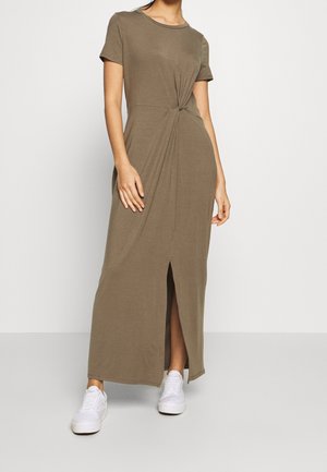 Oliwkowa sukienka do kolan z krótkimi rękawami, z przodu zawiązywana w supeł, z bocznym rozcięciem, noszona z białymi trampkami. Wykonana z miękkiego, elastycznego materiału.