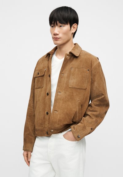 Καφέ σουέτ μπουφάν με γιακά, δύο τσέπες στο στήθος και κούμπωμα με κουμπιά. Διαθέτει κοντό σχέδιο και απαλό ύφασμα, φορεμένο πάνω από λευκό μπλουζάκι.