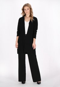 Cardigan lungo nero aperto davanti con maniche lunghe, abbinato a pantaloni neri a vita alta e gamba larga e tacchi neri. Tessuto a maglia testurizzata.