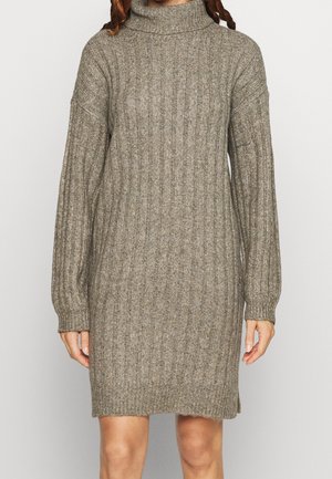 Woman wearing a knee-length, long-sleeve, ribbed knit taupe sweater dress with a high turtleneck collar.