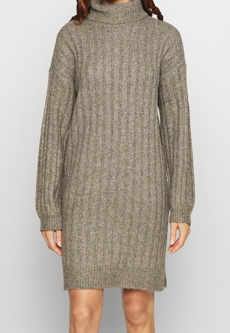 Kvinde iført en knælængde, langærmet, ribstrikket sweaterkjole i taupe med høj rullekrave.