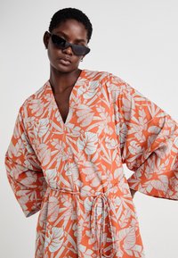 Robe portefeuille fleurie avec un fond orange et des motifs de plantes bleues, décolleté en V et manches larges, nouée à la taille avec une ceinture.