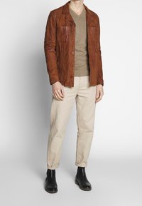 Giacca di pelle marrone con due tasche frontali, chiusura a bottoni e finitura testurizzata; abbinata a un maglione beige e pantaloni chinos crema. Stivali neri alla caviglia.