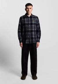 Camisa de franela a cuadros en negro y gris con cierre de botones, mangas largas y un pequeño logo amarillo. Combinada con pantalones anchos negros y zapatos marrones.
