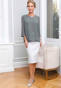 Graue Chiffonbluse mit rundem Ausschnitt und Dreiviertelärmeln, kombiniert mit einem taillierten weißen Bleistiftrock mit Blättermuster. Silberne spitz zulaufende Pumps.