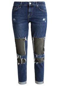 Modré džíny z poškozeného denimového materiálu s vyříznutými záplatami z olivově zelené látky, s rovnými nohavicemi a ohrnutým lemem s třepícími se okraji.